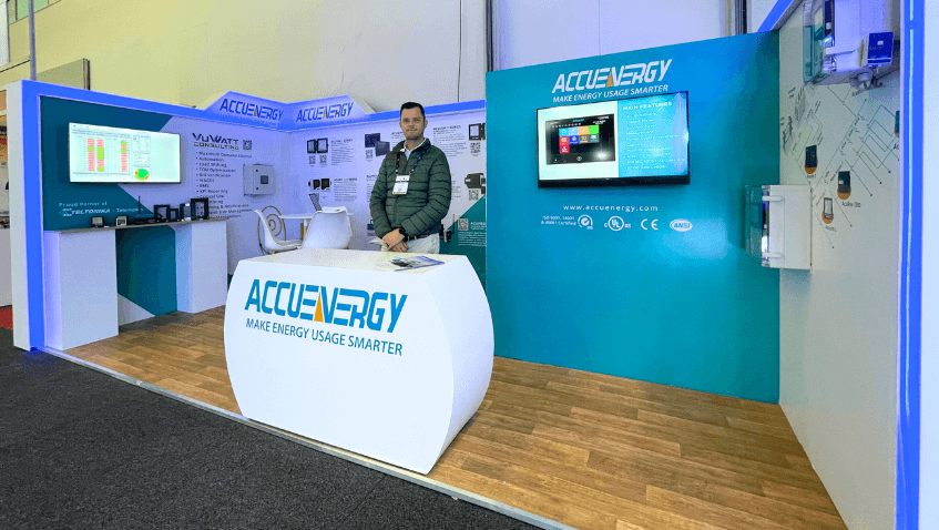 Accuenergy trade show booth featuring energy metering products, informational graphics, and a wall-mounted monitor at Enlit Africa 2025. Willie Botha, Business Development Manager, stands behind a branded reception counter, welcoming attendees.